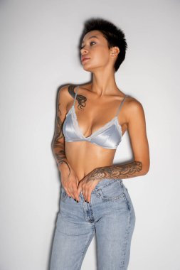 sexy tattooed woman in silk bra and jeans looking away on grey background