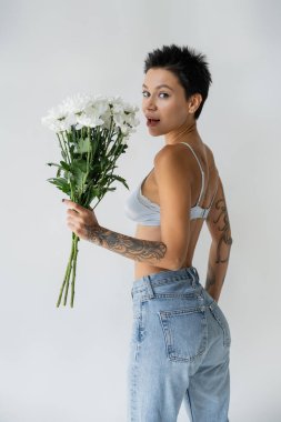 amazed woman in bralette and blue jeans holding bouquet of white flowers and looking at camera isolated on grey