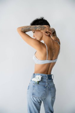tattooed woman in bra and jeans with white flowers in back pocket standing with hands behind head isolated on grey