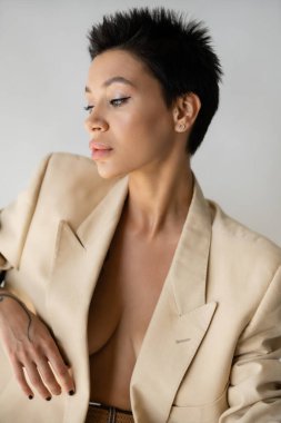 portrait of sexy brunette woman in beige blazer on shirtless body isolated on grey