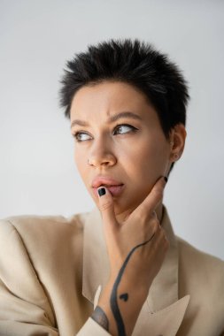 portrait of thoughtful woman in beige blazer touching lip and looking away isolated on grey