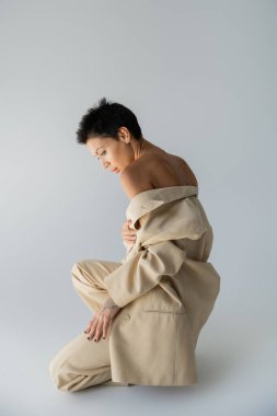 side view of sexy woman in beige suit posing with bare shoulders on grey background