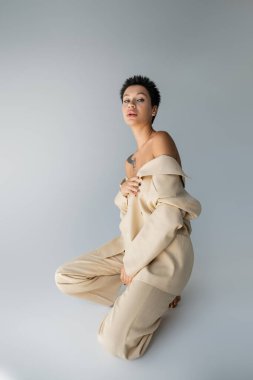 seductive brunette woman with bare shoulders posing in oversize suit on grey background