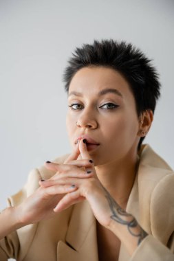 portrait of brunette woman with short hair and makeup touching lips and looking at camera isolated on grey
