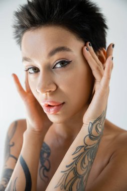 portrait of pretty tattooed woman with makeup touching face and looking at camera isolated on grey
