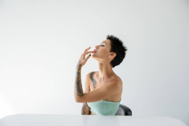 sexy tattooed woman touching lip while sitting near table and looking away on grey background clipart