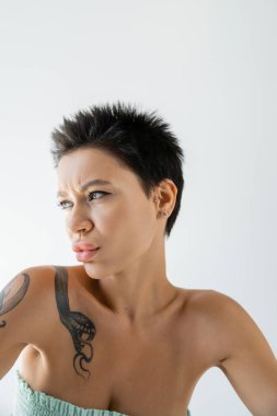 portrait of displeased tattooed woman in strapless top looking away isolated on grey