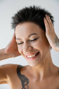 portrait of cheerful tattooed woman touching head while smiling with closed eyes isolated on grey