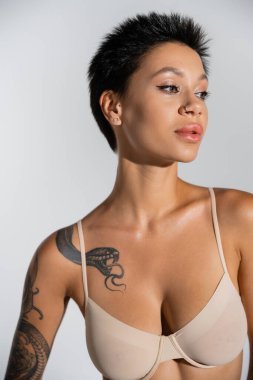 Dövmeli esmer kadın portresi. Makyajlı ve piercingli. Gri sütyenli seksi poz veriyor.