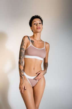 slender tattooed woman in bra and panties looking at camera on grey background