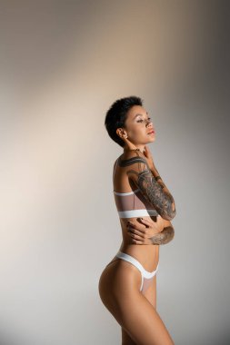 side view of sexy tattooed woman in underwear touching perfect body on grey background