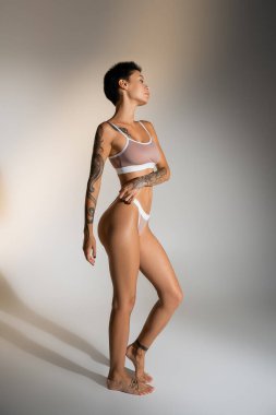 side view of slim brunette woman with tattooed body posing in lingerie on grey background