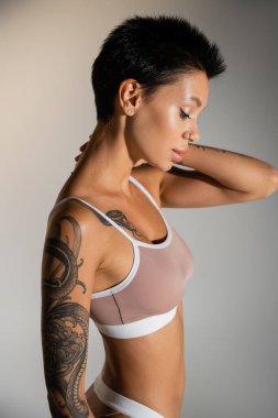 side view of brunette woman with tattooed body posing in bra with hand near neck on grey background