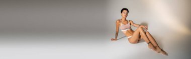 full length of fit woman with tattooed body posing in lingerie on grey background, banner clipart