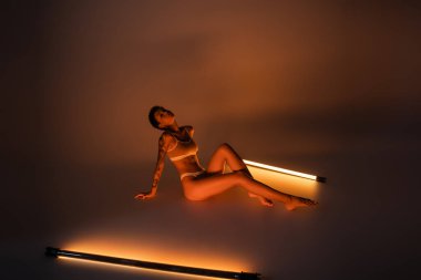 full length of seductive woman with slim tattooed body sitting in underwear near fluorescent lamps on dark background clipart