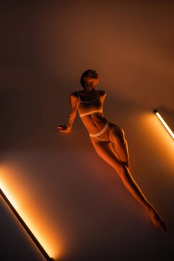 Yüksek açılı, iç çamaşırlı, siyah arka planda parlak lambaların yanında poz veren seksi bir kadın.