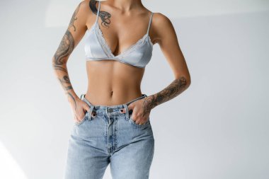 partial view of tattooed and slim woman in silk bralette holding hands in pockets of blue jeans on grey background