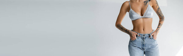 обрезанный вид стройная женщина в атласном бюстгальтере стоя с руками в карманах джинсов на сером фоне, баннер