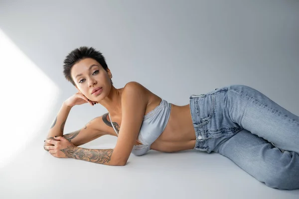 short haired and tattooed woman in blue jeans with bra lying on grey background