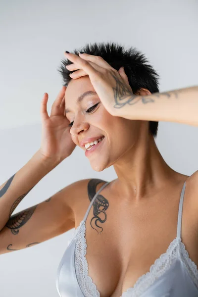 happy tattooed woman in blue bralette posing with hands near face isolated on grey
