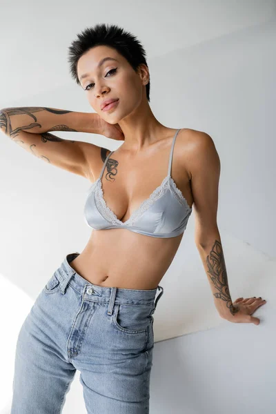 seductive tattooed woman in blue bra and jeans posing near cube with hand behind neck on grey background