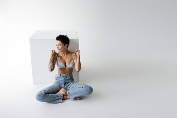 smiling tattooed woman touching straps of bra while sitting in blue jeans near cube on grey background