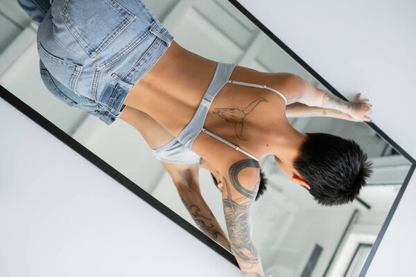tattooed short haired woman in bra and jeans lying on mirror on grey background