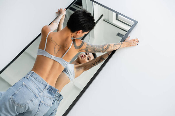high angle view of seductive tattooed woman in bra and jeans lying on mirror on grey background
