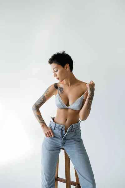 stock image tattooed woman in unzipped jeans and silk bra posing near high stool on grey background