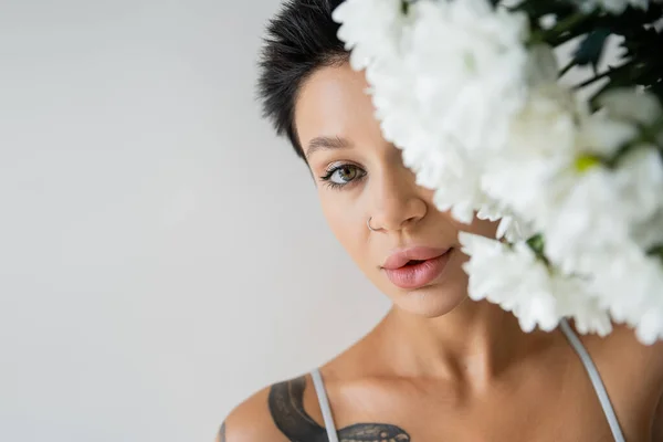 portrait of tattooed woman with makeup obscuring face with white flowers and looking at camera isolated on grey