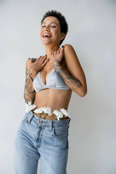 excited tattooed woman in silk bralette and jeans with white flowers laughing with closed eyes isolated on grey