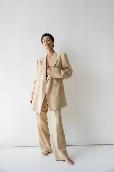 full length of barefoot woman in elegant oversize suit standing and looking at camera on grey background