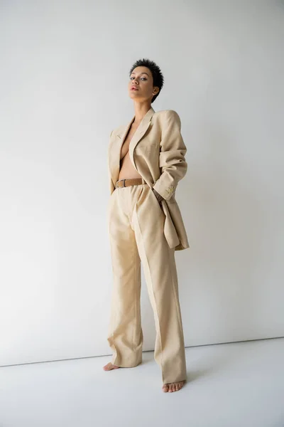 stock image full length of barefoot and shirtless woman in oversize pantsuit standing with hand in pocket on grey background