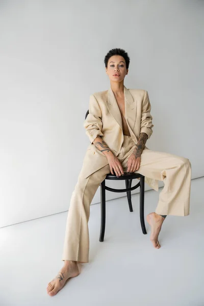 full length of barefoot and sexy woman in oversize suit posing on chair and looking at camera on grey background