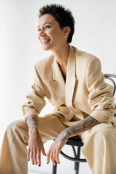 happy tattooed woman in fashionable pantsuit sitting on chair and looking away on grey background