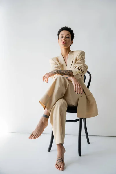 full length of tattooed barefoot woman in beige suit sitting on chair on grey background