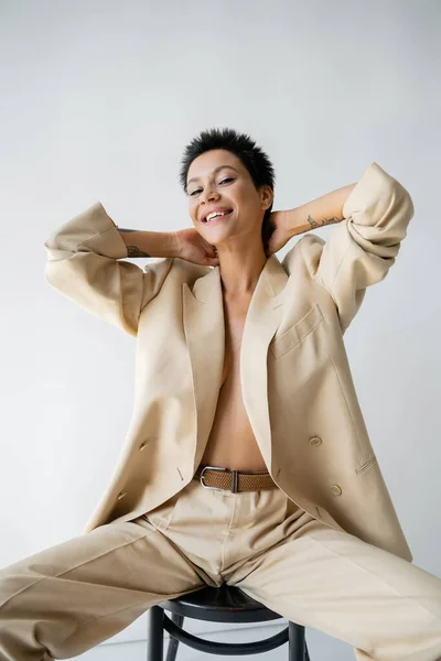happy shirtless woman wearing blazer on shirtless body and sitting on chair with hands behind neck on grey background