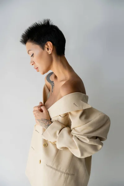 stock image side view of seductive tattooed woman with bare shoulders posing with oversize blazer on grey background