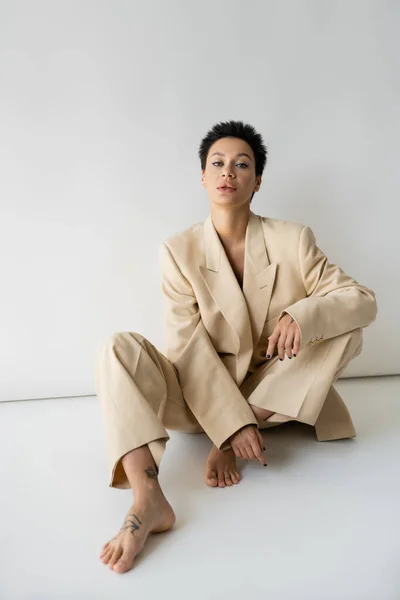 barefoot woman with short brunette hair looking at camera while sitting in beige suit on grey background