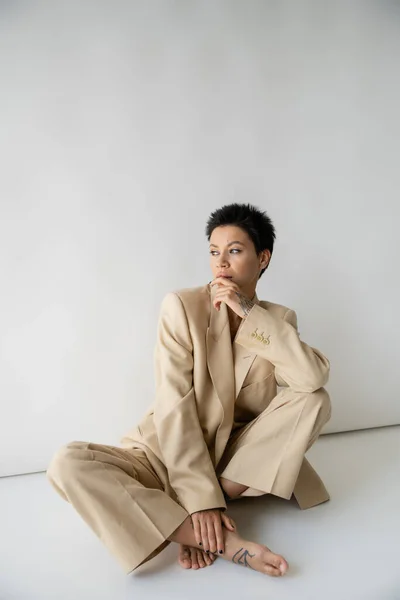 stock image pensive barefoot woman in beige suit sitting with crossed legs and looking away on grey background