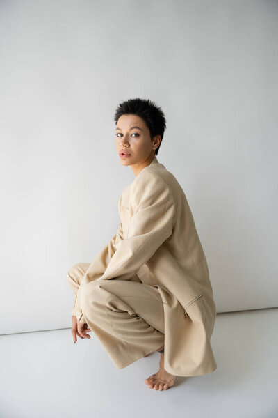 brunette woman in trendy suit sitting on haunches and looking at camera on grey background