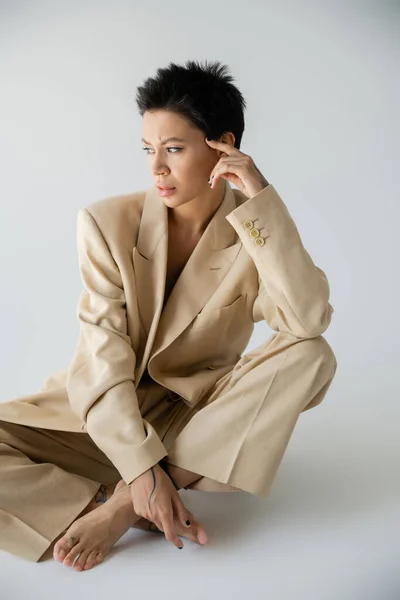thoughtful barefoot woman in stylish pantsuit sitting with crossed legs and looking away on grey background