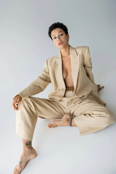 shirtless and barefoot woman in elegant pantsuit looking at camera while sitting on grey background