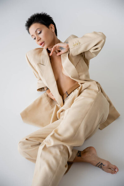 young and sexy woman in beige suit on shirtless body sitting on grey background