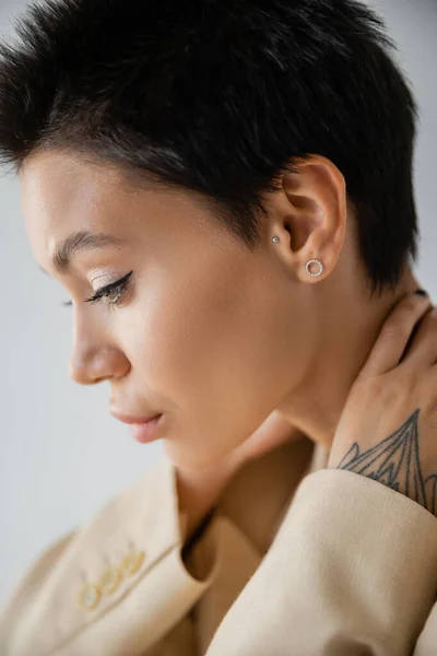 profile of young brunette woman with makeup and earrings posing with hands behind neck isolated on grey