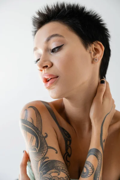portrait of sensual tattooed woman with bare shoulders posing with hand near neck isolated on grey