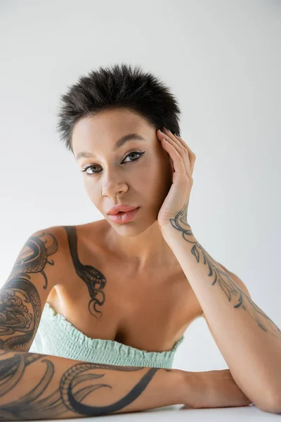 portrait of tattooed woman in strapless top posing with hand near face isolated on grey
