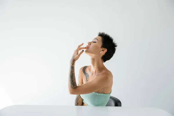 sexy tattooed woman touching lip while sitting near table and looking away on grey background