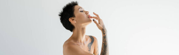 Вид сбоку татуированной брюнетки с голыми плечами, касающимися губ и отводящими взгляд на сером фоне, баннер