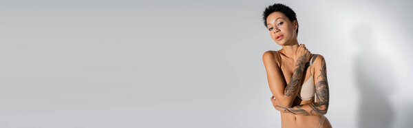 татуированная брюнетка в лифчике, смотрящая на камеру на сером фоне, баннер